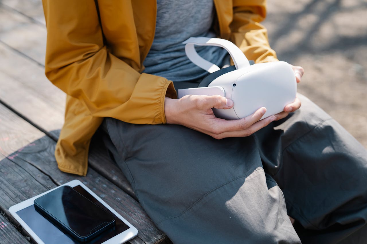From above of crop unrecognizable person sitting on wooden bench near smartphone and tablet while holding VR headset in hands